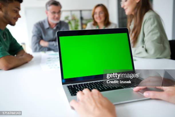 manos recortadas de una mujer de negocios sobre una computadora portátil con una pantalla verde en blanco contra colegas en la oficina - portatil en oficina subjetivo pantalla fotografías e imágenes de stock