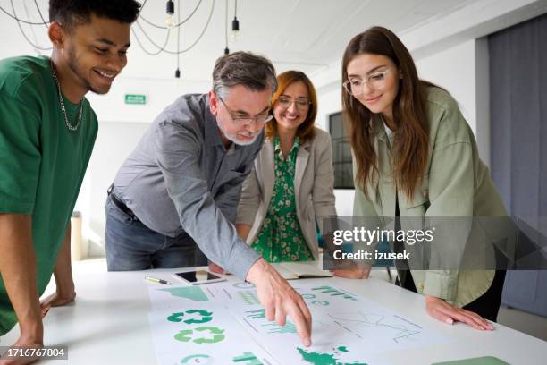 mature businessman pointing at chart while brainstorming with colleagues in office during meeting - duurzaam bedrijf stockfoto's en -beelden