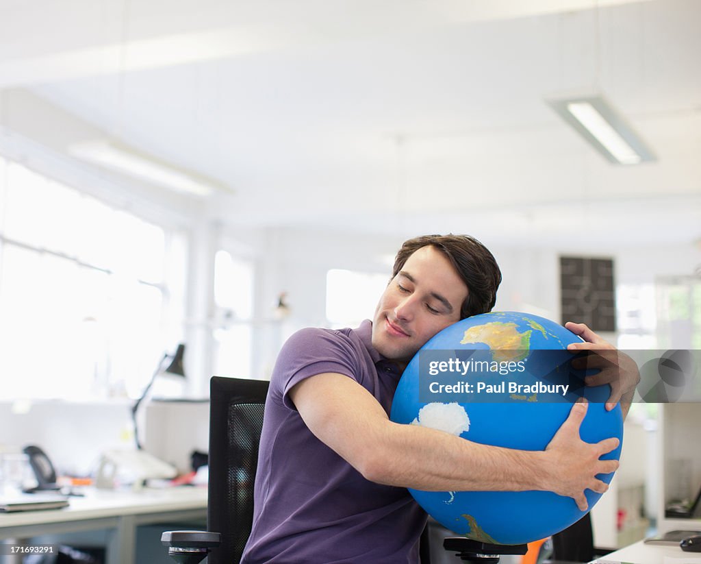 Businessman with eyes closed hugging globe at desk in office