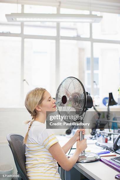 femme souriante, assis en face de fan dans le bureau - ventilateur électrique photos et images de collection