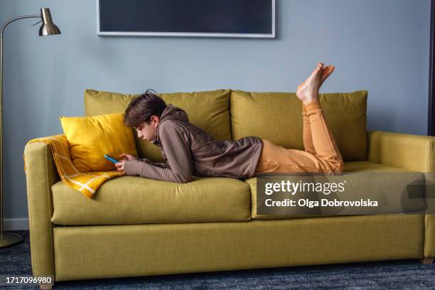 a teenage boy lying down on a sofa, resting on his elbows and looking through his mobile phone - sole of foot stock pictures, royalty-free photos & images