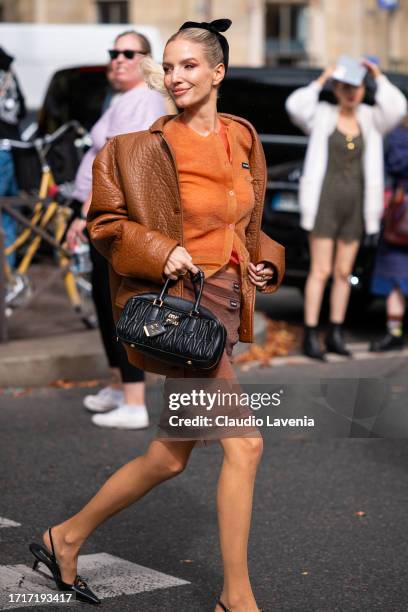 Leonie Hanne wears an orange Miu Miu cardigan, brown leather jacket, brown sheer Miu Miu miini skirt, black Miu Miu bag and black kitten heels,...