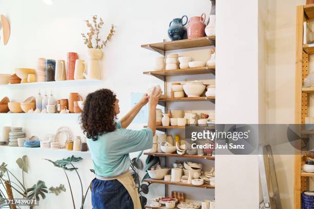 mujer alfarera irreconocible arreglando tazas de cerámica en el estante del taller a la luz del día - ceramics fotografías e imágenes de stock