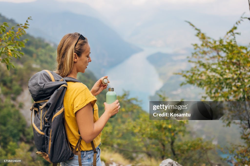 Una mujer cierra una botella de agua de acero inoxidable mientras está de pie en la cima de una montaña y disfruta de la naturaleza que la rodea