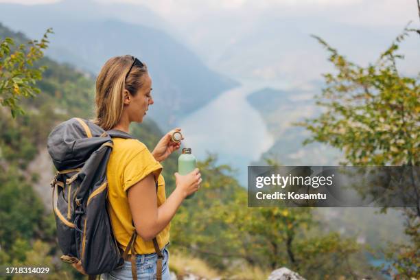 a woman closes a stainless steel water bottle while standing on top of a mountain and enjoying the nature that surrounds her - sustainable tourism stock pictures, royalty-free photos & images