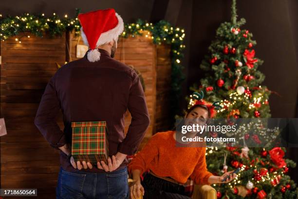rückansicht eines jungen mannes, der ein weihnachtsgeschenk hinter seinem rücken hält und seine freundin überraschen will - nikolausmütze stock-fotos und bilder