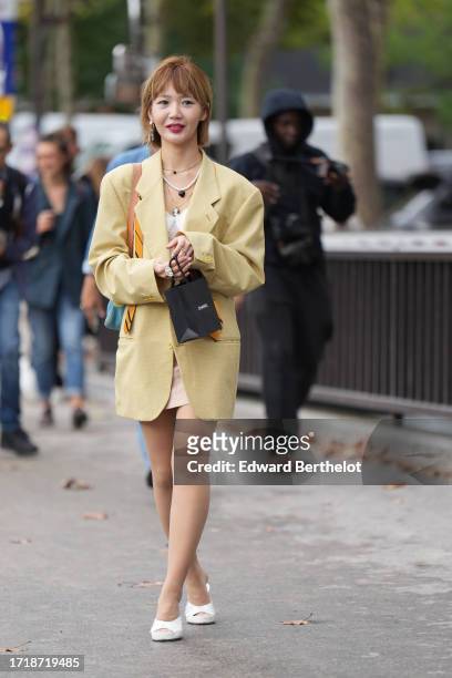 Guest wears earrings, a necklace, a yellow long oversized pastel color yellow blazer jacket worn as a dress, white shoes, outside Chanel, during the...