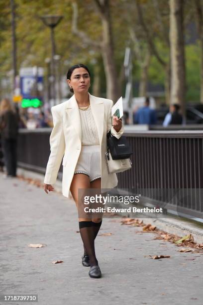 Guest wears a golden necklace, a white oversized blazer jacket, a white knit pullover made of wool, mini shorts, black leather shoes from Prada,...