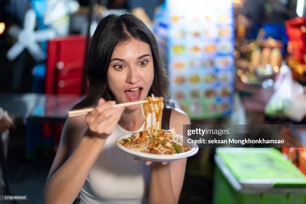 Young woman eating Pad Thai while traveling on Khao San Road at night in Bangkok, Thailand