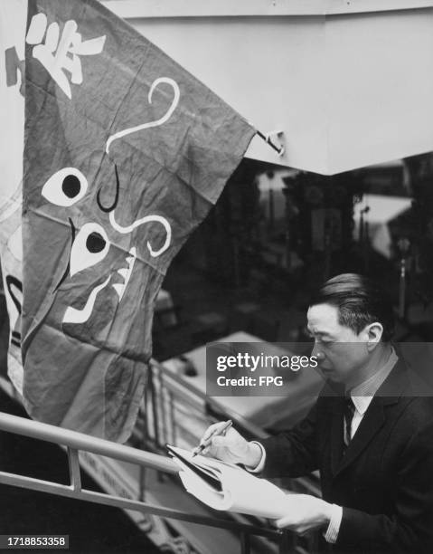 Chinese-American artist Dong Kingman writing in a notebook as he stands beneath a flag featuring an abstract design and a Chinese character, United...