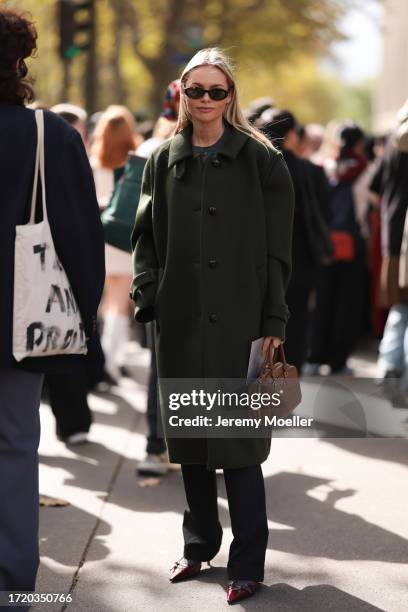 Claire Rose Cliteur is seen wearing a long olive green wool coat, black trousers, red patent shoes, black brown oval sunglasses and a brown leather...