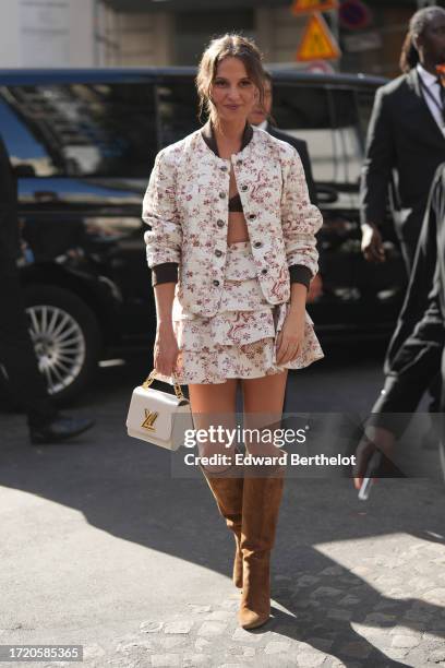 Alicia Vikander wears a white and pink floral print jacket, a gathered and ruffled mini skirt, black bras, a white LV bag, knee-high brown suede...