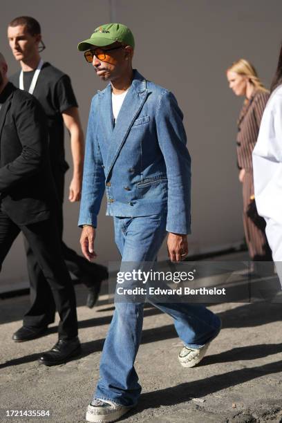 Pharrell Williams wears a green cap hat, a t-shirt, a blue denim double breasted jacket, flared jeans / denim pants, sneakers, outside Louis Vuitton,...