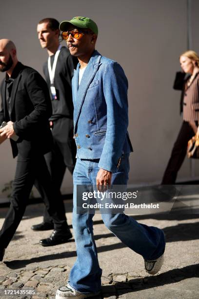 Pharrell Williams wears a green cap hat, a t-shirt, a blue denim double breasted jacket, flared jeans / denim pants, sneakers, outside Louis Vuitton,...