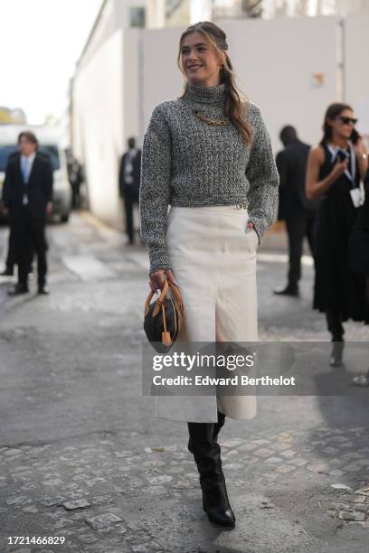 Zita D'Hauteville wears a gray wool turtleneck pullover, a golden necklace, a white slit midi skirt, knee high leather boots, a brown Vuitton bag,...