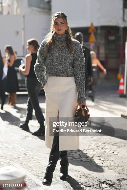 Zita D'Hauteville wears a gray wool turtleneck pullover, a golden necklace, a white slit midi skirt, knee high leather boots, a brown Vuitton bag,...
