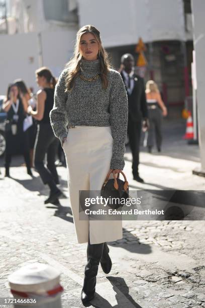 Zita D'Hauteville wears a gray wool turtleneck pullover, a golden necklace, a white slit midi skirt, knee high leather boots, a brown Vuitton bag,...