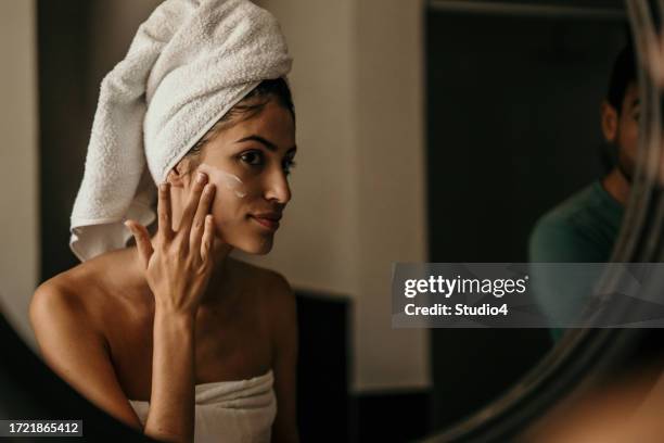 eine junge frau trägt eine feuchtigkeitscreme auf ihr gesicht auf, ihr spiegelbild fängt ihre hingabe zur selbstfürsorge ein - haushaltsarmatur stock-fotos und bilder