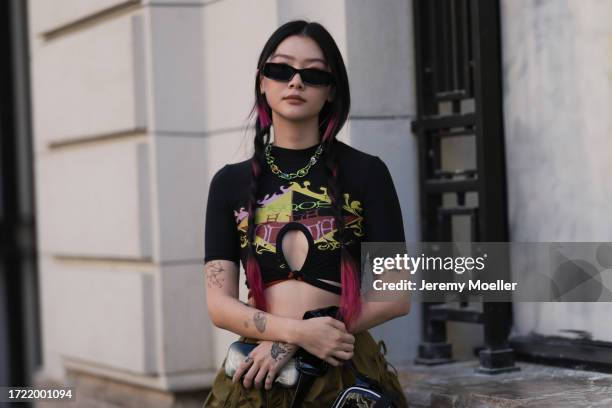 Katie Qian is seen wearing green wide trousers, a black Ottolinger x Puma bag, a cropped black shirt with colorful writings on it, black shades and a...