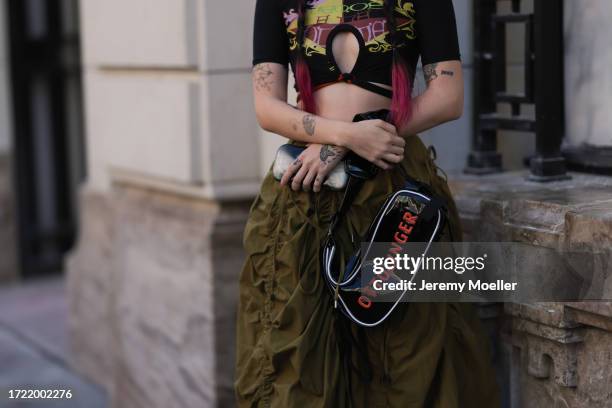 Katie Qian is seen wearing green wide trousers, a black Ottolinger x Puma bag, a cropped black shirt with colorful writings on it, black shades and a...