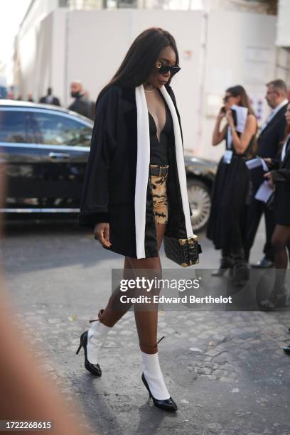 Venus Williams wears sunglasses, a black jacket with white stripes on the lapels, a low-neck top, a belt, military camo shorts, white socks pointed...