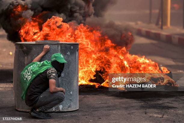 Masked Palestinian protester takes cover near flaming tires during clashes with Israeli forces following a rally in solidarity with Gaza by...
