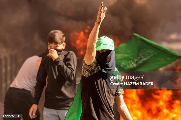 Masked Palestinian protester gestures while walking near flaming tires during clashes with Israeli forces following a rally in solidarity with Gaza...