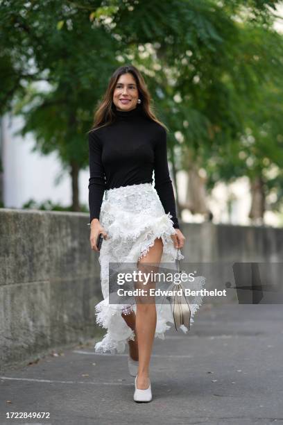 Belen Hostalet wears earrings, a black turtleneck pullover, a white lace floral embroidery asymmetric skirt, a cream color bag, white shoes, outside...