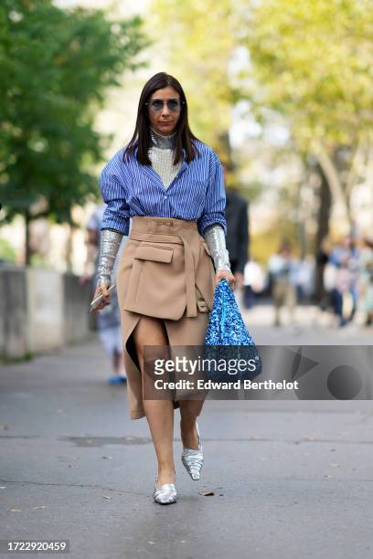 Guest wears sunglasses, silver sequined turtleneck shiny top, a blue striped shirt, a beige asymmetric thick slit skirt, a blue sequined shiny bag,...