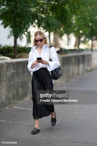 Guest wears sunglasses, a necklace, a white shirt, a black lustrous skirt, black loafers shoes, a bag, outside Zimmermann, during the Womenswear...