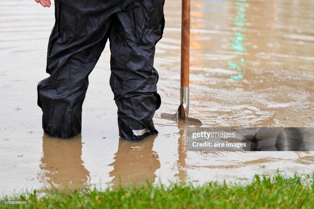 Rear view of a person standing in the street flooded with water trying to unclog the sewer with a tool