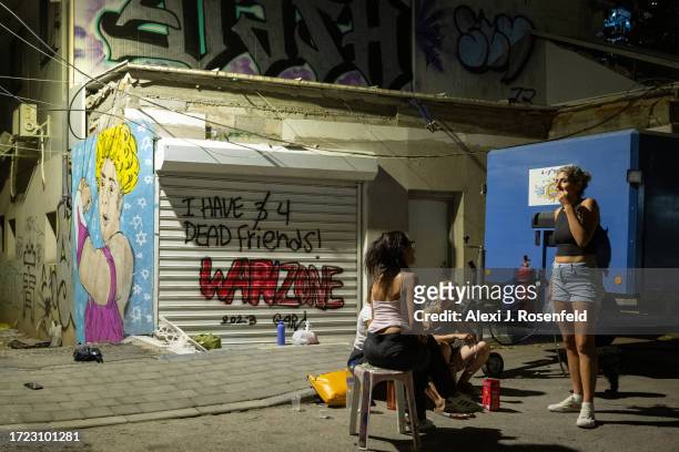 People sit on a street near new graffiti that reads, I have 4 dead friends, warzone, and an drawing of Rachel, who was held by Hamas militants for 20...