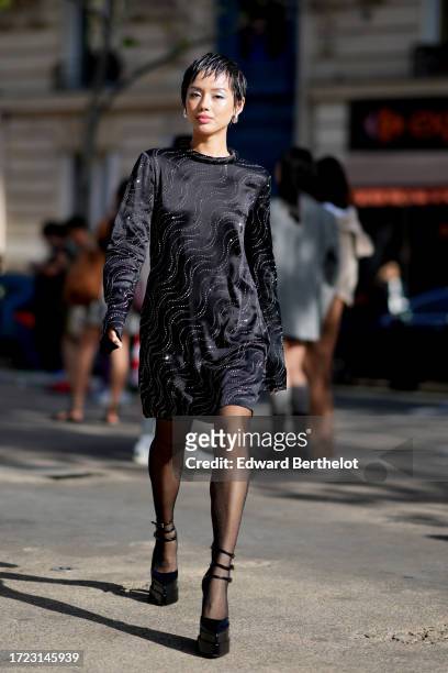 Khanh Linh wears earrings, a black lustrous silk mini dress with long sleeves and printed geometric patterns, outside Stella McCartney, during the...