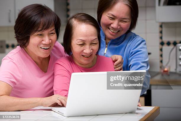 asian adult women laughing and smiling, using laptop in kitchen - only mature women stock pictures, royalty-free photos & images