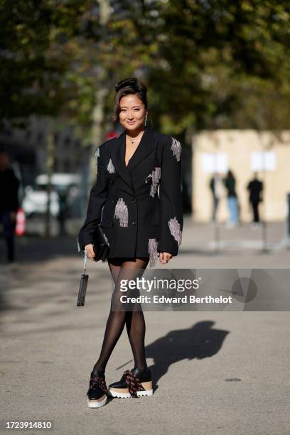 Ashley Park wears earrings, a black oversized blazer jacket with bejeweled attached fringes, black tights, black leather shoes with brown shoelace...