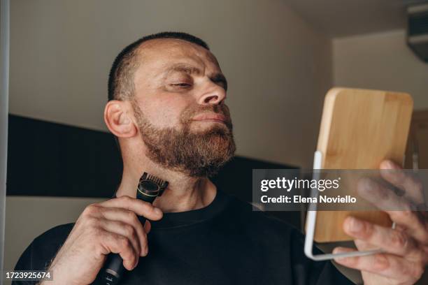 a man shaves his beard with an electric razor and looks in the mirror, men's skin and facial care - electric razor stock pictures, royalty-free photos & images
