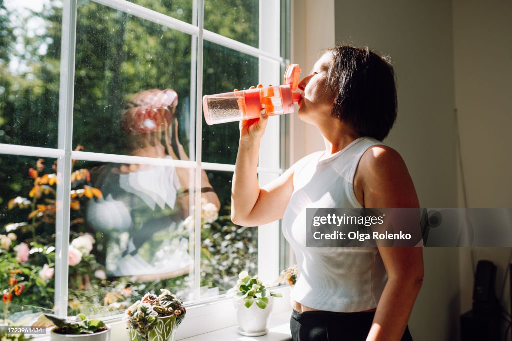 Replenishment of water balance. Mature woman drink water after home exercising staying against large window