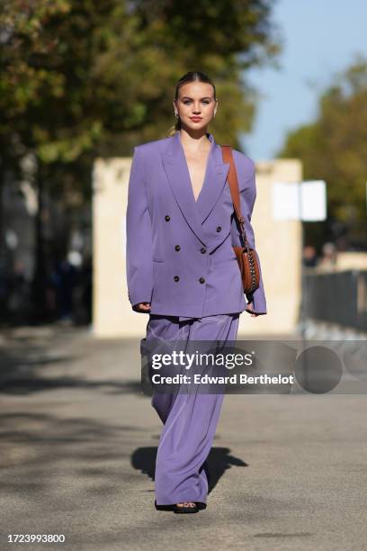 Guest wears earrings, a purple / mauve pastel oversized blazer jacket, a brown leather bag, matching flared pants, outside Stella McCartney, during...