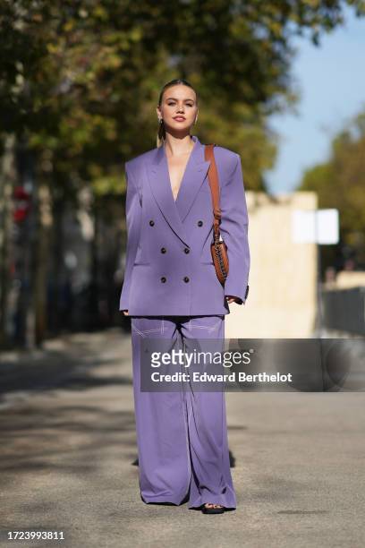 Guest wears earrings, a purple / mauve pastel oversized blazer jacket, a brown leather bag, matching flared pants, outside Stella McCartney, during...