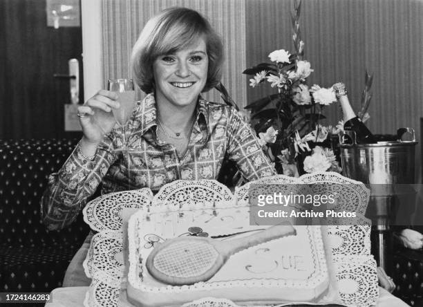 British tennis player Sue Barker raises a glass of champagne as she smiles behind her 21st birthday cake, which features a tennis racquet, April 1977.