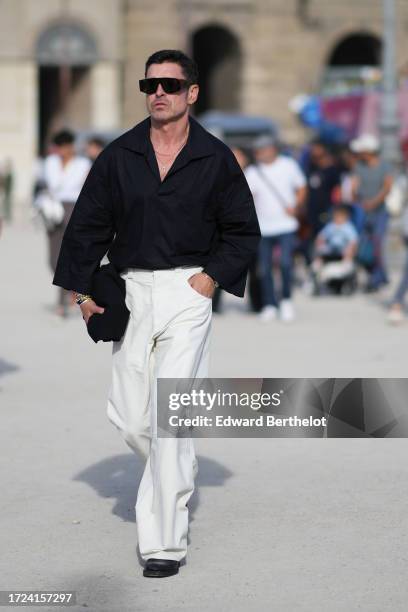 Alex Badia wears sunglasses, a black shirt with flared sleeves, white flared pats, black shoes, outside Casablanca, during the Womenswear...