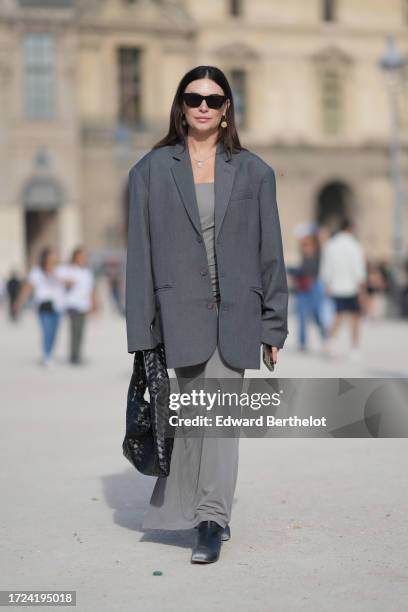 Guest wears sunglasses, a gray oversized blazer jacket, a gray long dress, a black woven leather bag, boots, outside Casablanca, during the...