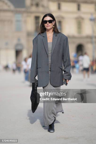 Guest wears sunglasses, a gray oversized blazer jacket, a gray long dress, a black woven leather bag, boots, outside Casablanca, during the...