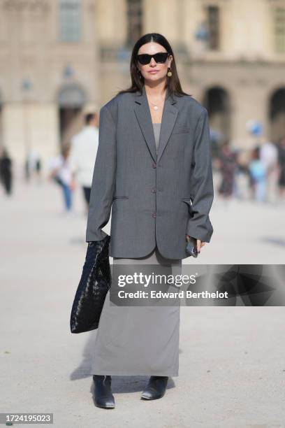 Guest wears sunglasses, a gray oversized blazer jacket, a gray long dress, a black woven leather bag, boots, outside Casablanca, during the...