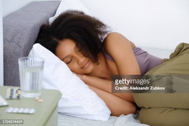 attractive sleeping dark-skinned woman or girl in the bedroom. a glass of water, pills, medicines and medicines on the bedside table next to the bed. the concept of lack of sleep, fatigue, lack of melatonin. treatment and prevention of insomnia. - melatonin fotografías e imágenes de stock