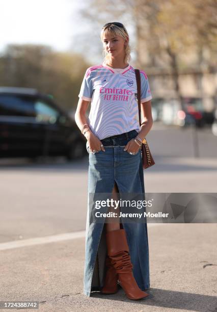 Mia Regan is seen wearing a blue, pink sports T-shirt, a blue jeans maxi skirt with slit, high brown leather boots, a brown crossbody handbag and...