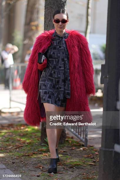 Andreea Cristea wears sunglasses, a red long faux fur coat, a gray short dress with printed geometric patterns, leather shoes, outside Akris, during...