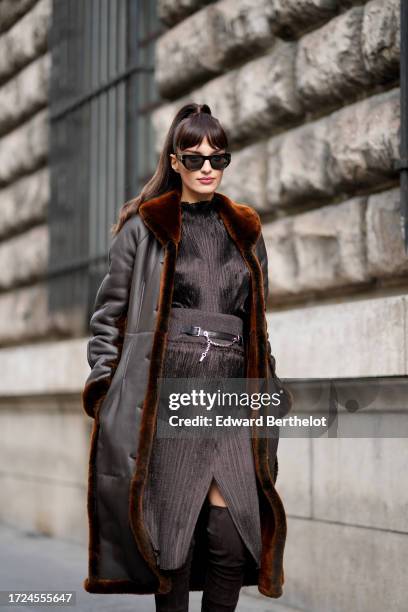 Gabrielle Caunesil wears sunglasses, a brown long leather aviator jacket with fluffy inner lining, a brown ruffled ribbed satin dress with large...