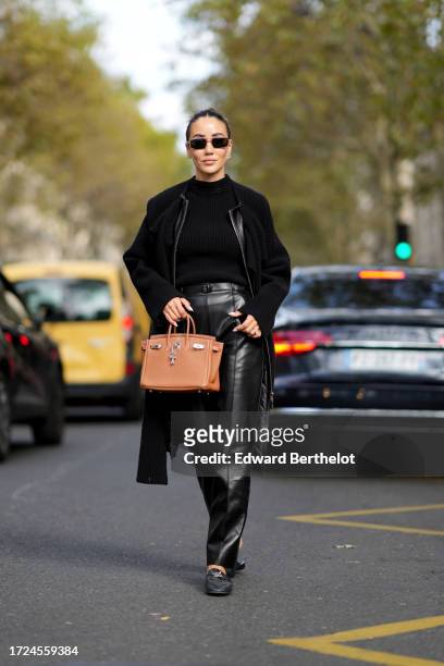 Tamara Kalinic wears sunglasses, a black wool ribbed pullover, a long coat, a brown Hermes bag, a leather jacket, leather black pants, loafers shoes,...