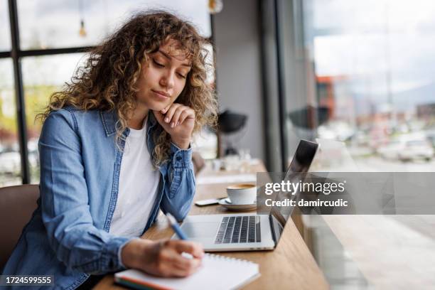 young woman working on laptop and taking notes at a cafe - werk zoeken stockfoto's en -beelden
